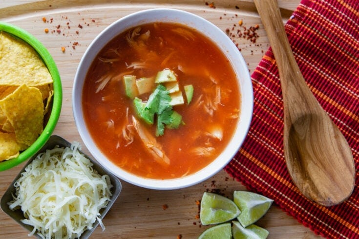Chicken tortilla soup with tortilla chips, cheese, lime garnish and wooden spoon.