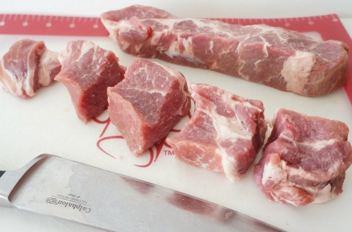 Sliced chunks of pork and whole cut of pork arranged on a white cutting board with a chef's knife.