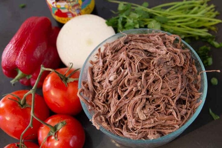 Glass bowl of shredded beef centered to the right and flanked by vegetables.