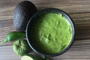 Bowl of green salsa accompanied by avocado and peppers.