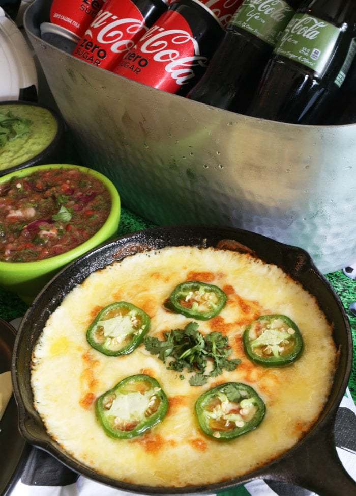 Queso fundido topped with jalapeno slices in a cast iron pan alongside small bowls of dip.