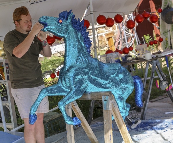 Artist painting the muzzle region of a glittering blue horse sculpture at Camp Christmas.