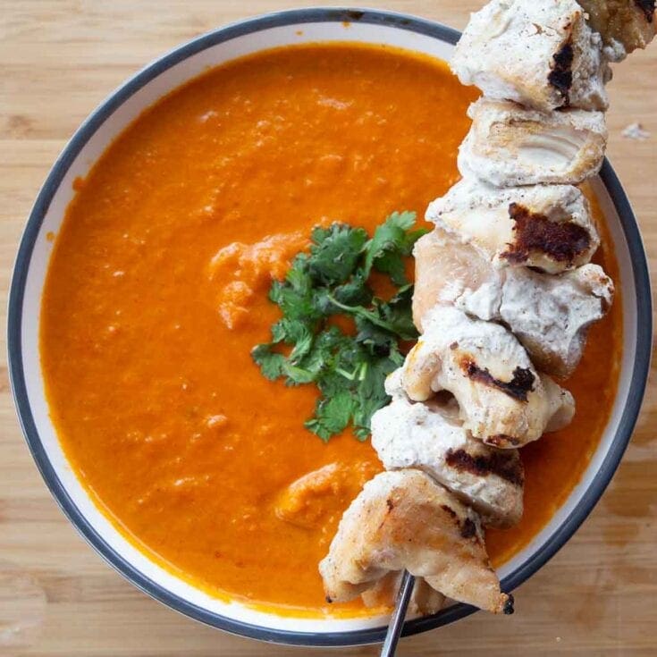 Tandoori chicken skewer lying diagonally across a cilantro-topped bowl of soup.