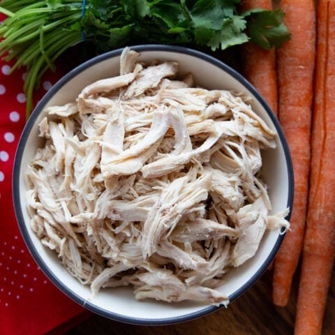 Bowl of shredded chicken flanked by a bunch of cilantro, carrots, a cloth, and a red pepper.
