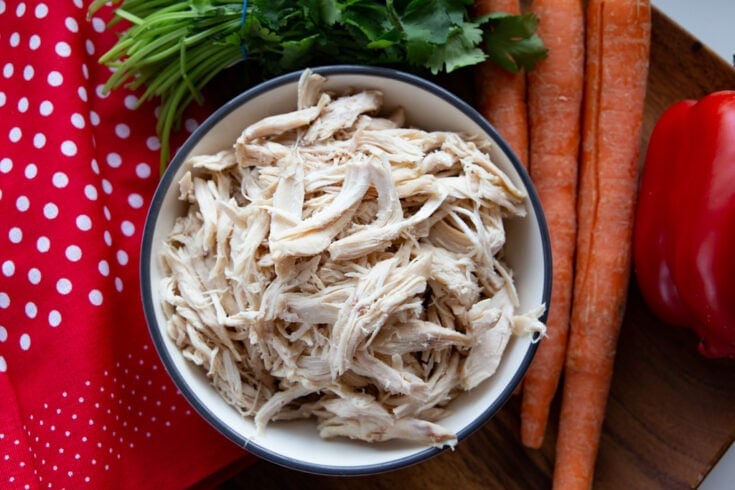 Bowl of shredded chicken flanked by a bunch of cilantro, carrots, a cloth, and a red pepper.