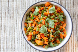 Bowl full of diced carrots and green beans on a white woven tablecloth.